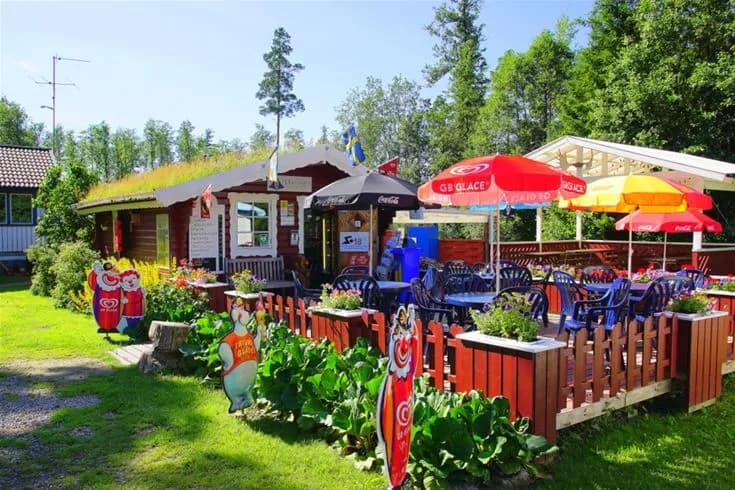 Alt-text: En lummig campingplats med gröna växter och välskötta gräsmattor på Otterbergets bad och camping. I bakgrunden finns en trädgård med parasoller som ger skugga, samt ett utomhusbord för avkoppling.