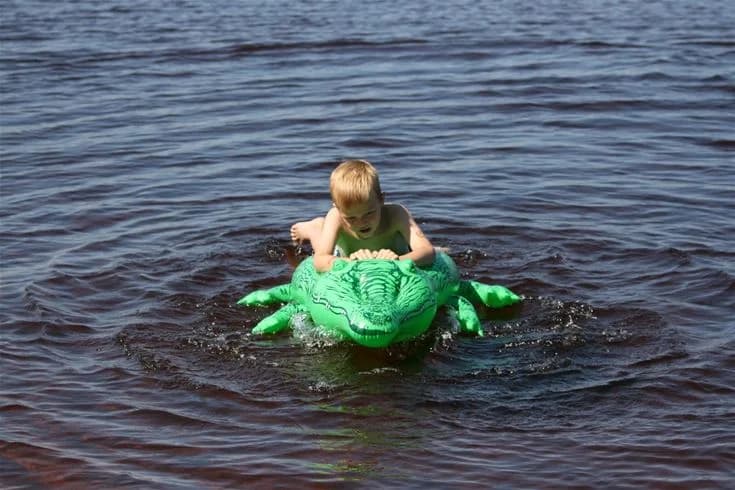 En grupp barn leker och har kul vid en sjö på Otterbergets bad och camping, omgivna av idyllisk natur och semesterstämning.