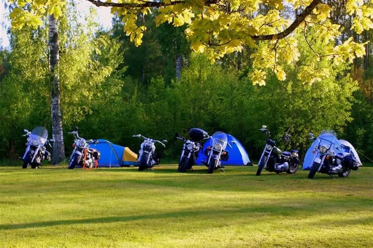 En solig dag på Otterbergets bad och camping. I förgrunden syns ett tält uppställt på en grön gräsmatta, omgivet av höga träd. Himlen är klarblå och platsen erbjuder en rofylld miljö för friluftsliv och rekreation.