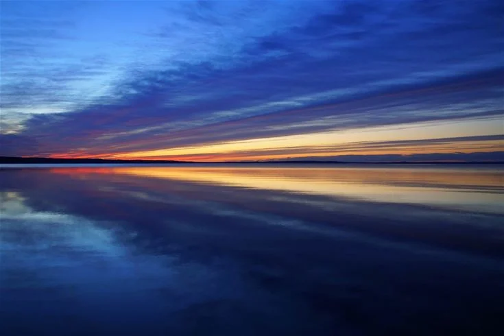 En stilla sjö vid Otterbergets bad och camping, med en blå himmel och vita moln som speglas i vattnet. Solnedgångens ljus färgar horisonten i mjuka orange och rosa toner.