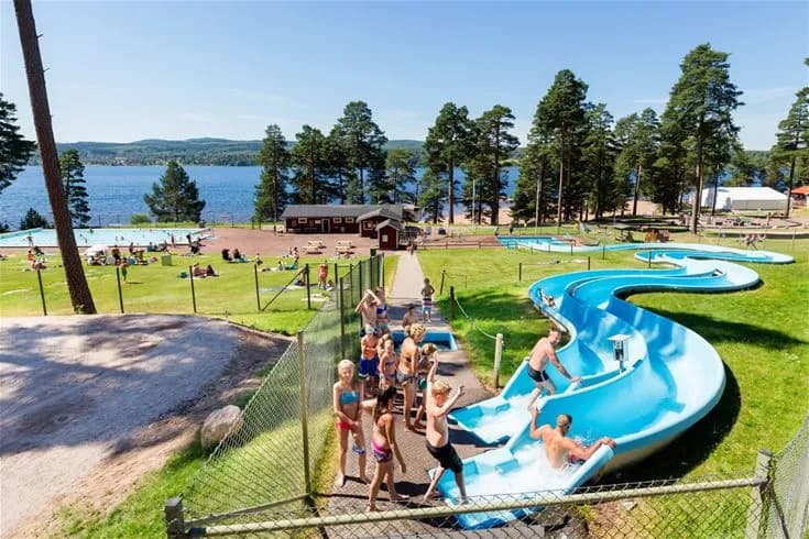 En solig dag på Leksand Resort med glada människor som njuter av poolen och de olika sommaraktiviteterna. Färgglada badkläder syns kring en stor utomhuspool, omgiven av grönskande gräsmattor och parker, vilket skapar en inbjudande semesteratmosfär.