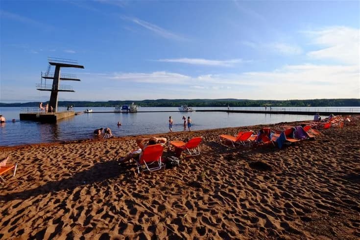En solig sommardag vid Leksand Resort, där människor njuter av semestern vid stranden. Vita cumulusmoln pryder den blå himlen, medan besökare kopplar av vid bryggan och engagerar sig i fritidsaktiviteter.