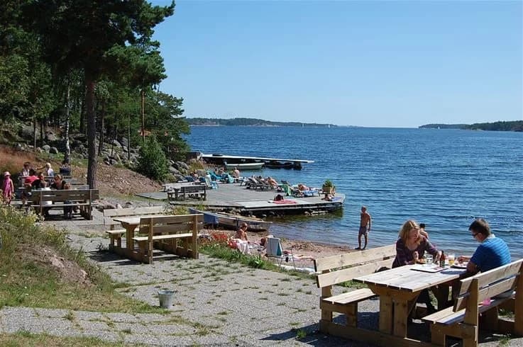 En solig sommardag vid Täntö havsbad & camping, med människor som njuter av sin semester. Utemöbler står uppställda vid kustlinjen, där besökare solar och kopplar av nära sjöns strand.