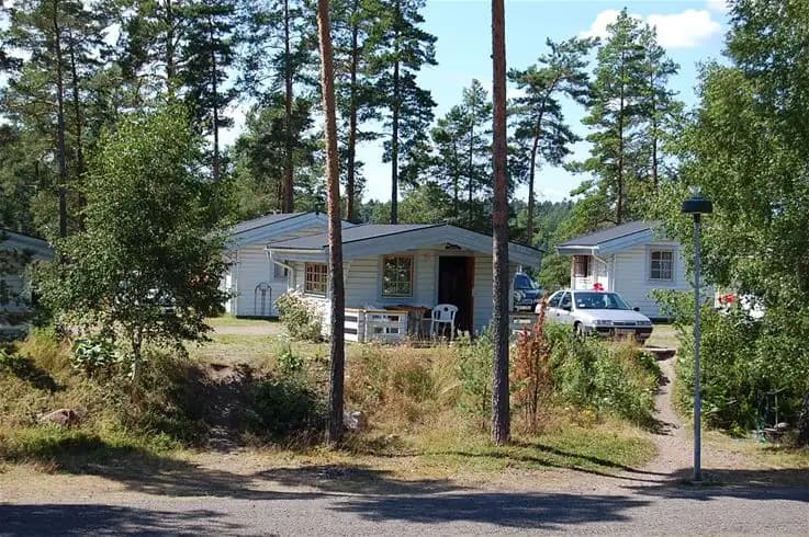 En lantlig stuga omgiven av träd och buskar på Tättö havsbad & camping. I förgrunden syns en välskött trädgård med gröna växter.
