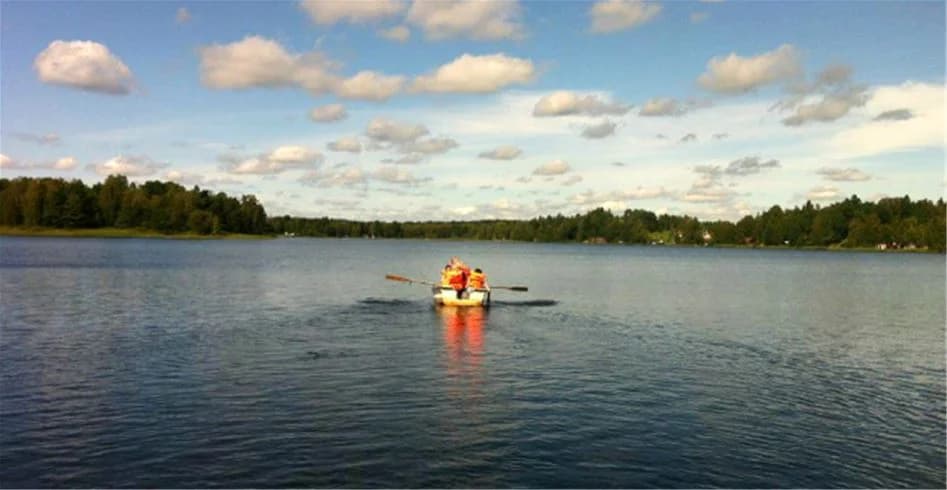 En person paddlar kajak på en lugn vattenyta under molnig himmel vid Eksjö Camping & Konferens. I bakgrunden syns andra båtar och en naturskön omgivning som inbjuder till friluftsaktiviteter.