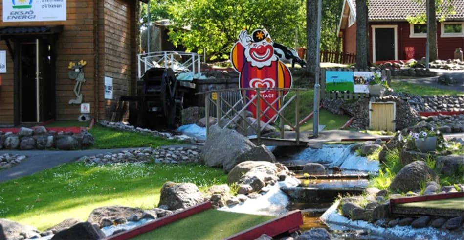 Alt-text: En bild av Eksjö Camping & Konferens som visar en mini-golfbana och en skylt med campingens logotyp, omgiven av grönskande parkmiljö.