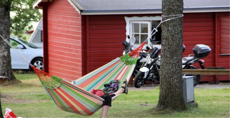 En solig campingplats med gröna gräsmattor och skuggiga områden, där en hängmatta hänger mellan träden, inbjudande för avkoppling och rekreation. Perfekt för semester och fritidsaktiviteter.