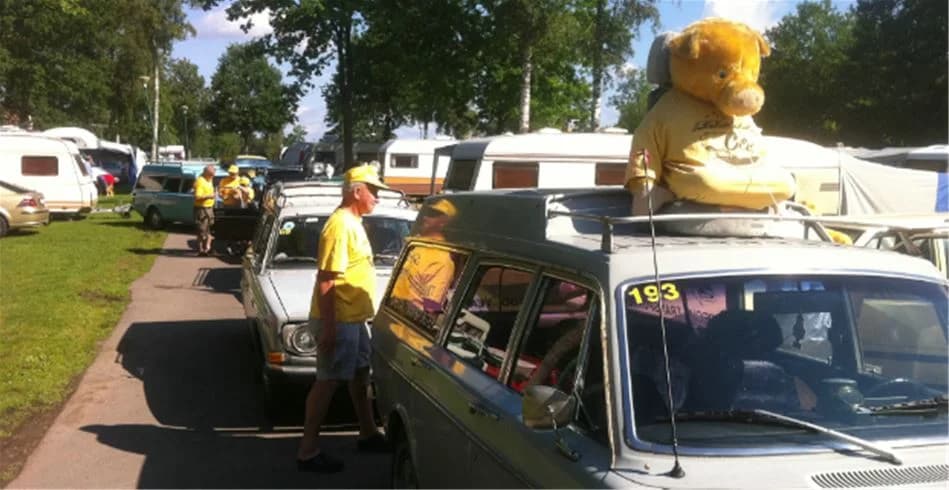 En klassisk familjebil med takräcke är parkerad vid sidan av en campingplats. I bakgrunden syns ett staket. 