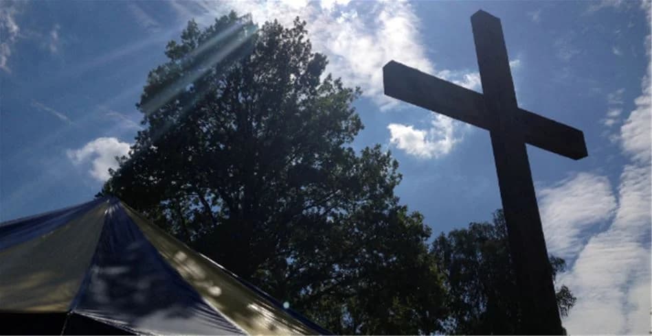 Alt-text: En solig morgon vid Eksjö Camping & Konferens där solens strålar skapar linsöverstrålning genom trädkronorna, och krucifixet står i kontrast mot den natursköna bakgrunden.