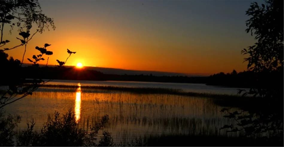 En vy över en stilla vattenyta vid Eksjö Camping & Konferens, där en vacker solnedgång färgar himlen i orange och rosa toner.
