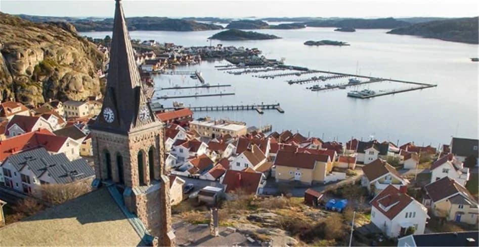 Vy över Fjällbacka Camping, med en stad i bakgrunden, inkluderar en kyrkspira och tak från bostadsområden, omgiven av kust och naturlandskap.