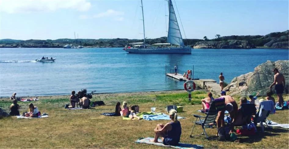 Vy över Fjällbacka camping med fokus på kusten. Människor njuter av fritidsaktiviteter vid havet. En båt syns vid strandlinjen och området är perfekt för en picknick och avkoppling.