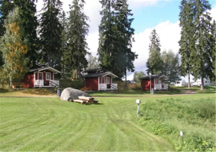 En grönskande campingplats med gräs, träd och små stugor, omgiven av en lummig landskapsmiljö.