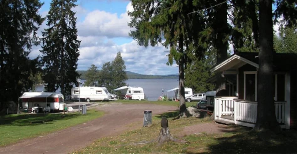 Bild av Munkebergs Camping med en sjö i förgrunden, omgiven av grönskande landskap. I bakgrunden skymtas några husvagnar och campingplatser vid sjöns strand.