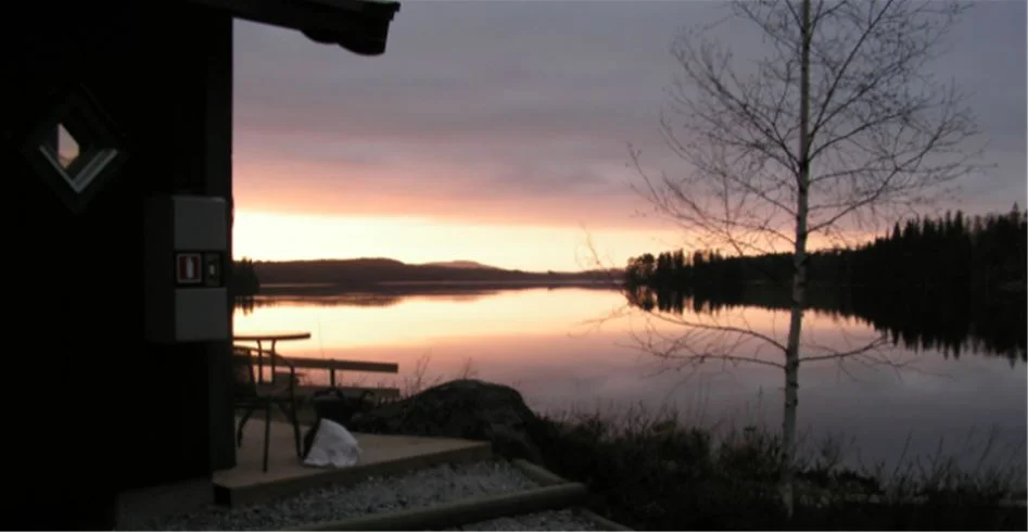 En lugn sjö med spegelblank yta omgiven av träd vid skymning, vars orange och rosa efterglöd reflekteras i vattnet, på Munkebergs Camping.