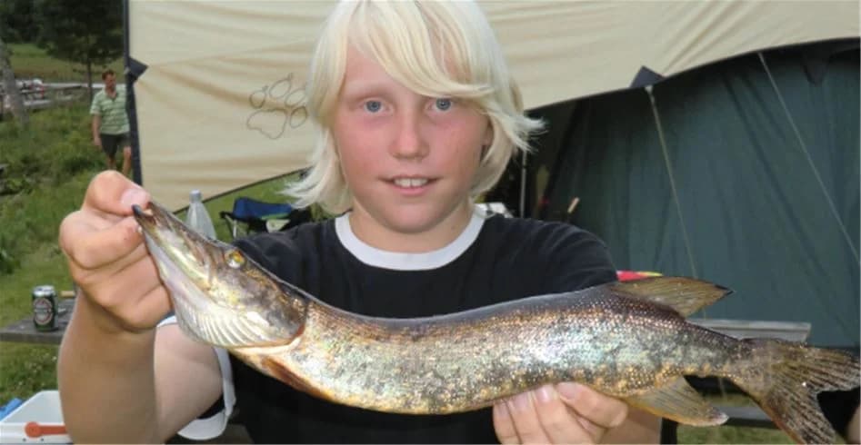 En glatt leende fiskare håller upp en stor gädda vid strandkanten på Munkebergs camping, omgivning med klart vatten och gröna buskar i bakgrunden.