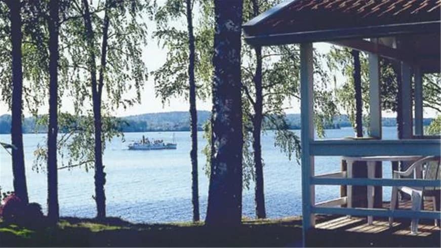 En naturskön campingplats vid vattnet, med skuggande träd längs strandkanten och en paviljong i bakgrunden.