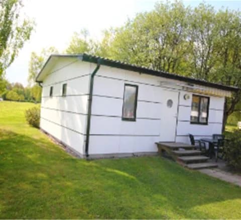 En liten stuga med rött tak och vita väggar står i en grön trädgård med välskött gräsmatta. I bakgrunden syns ytterligare byggnader och en husvagn på Gustavsvik Resort.