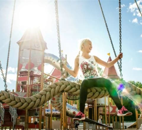 En solig dag på Gustavsvik Resort visar barn som leker glatt på en lekplats med modern lekutrustning. Ett barn i sneakers syns i förgrunden, medan andra roas av att använda olika delar av lekplatsen. En känsla av glädje och semester sprider sig i luften.