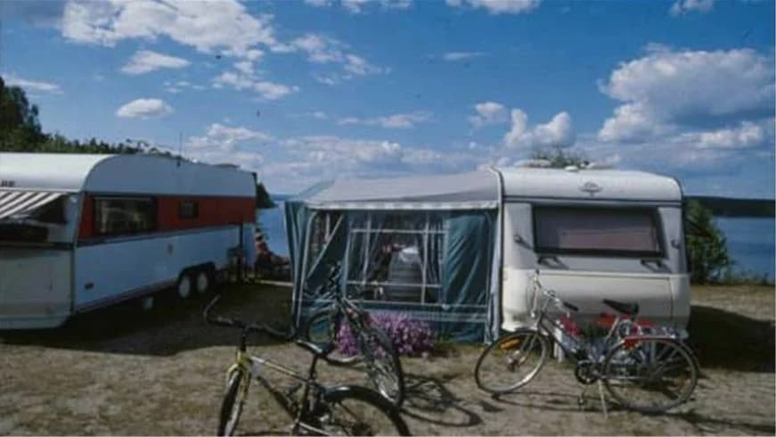 En campingplats med husvagnar, husbilar och tält på grön mark, där ett par klassiska bilar också står parkerade.