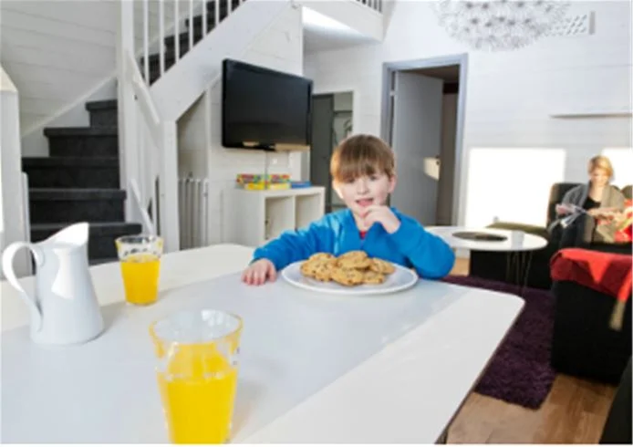En familj med ett litet barn sitter vid ett matbord i ett modernt kök på Skellefteå camping. Rummet är hemtrevligt inrett med möbler och köksutrustning. En ljus och trivsam atmosfär avspeglar sig i den gemensamma måltiden.