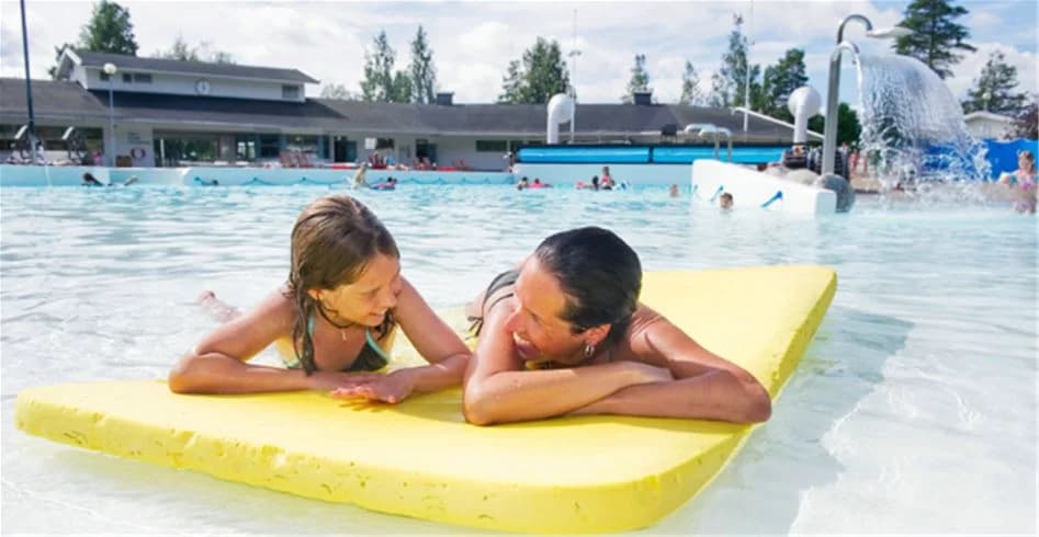 En livlig och glad scen vid Skellefteå camping, med barn och vuxna som njuter av sol och bad runt en stor swimmingpool. Personer i badkläder leker och kopplar av vid vattnet, omgivna av semesterkänsla och glada ansikten på ett populärt besöksmål.