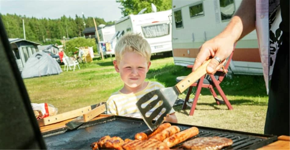 En familj grillar mat på en utomhusgrill på Skellefteå Camping, medan ett barn leker i bakgrunden.