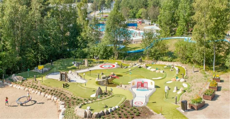 En solig dag på Skellefteå camping med ett välskött minigolfområde omgivet av gröna träd och lekande barn.