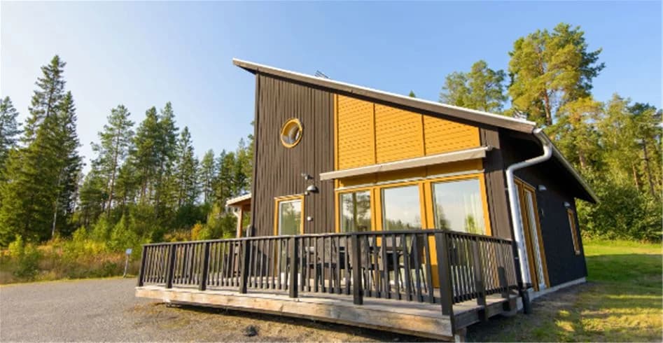 Alt-text: En mysig stuga i trä med veranda, belägen på Skellefteå camping. Stugan har klassisk arkitektur med träpanel och ett brunt sadeltak, omgiven av grönska.
