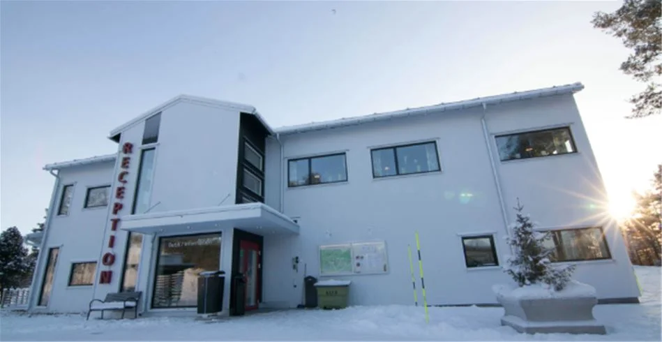 En snötäckt stuga står vid Skellefteå camping, omgiven av tjockt snötäcke. En fyrhjulsdriven bil är parkerad nära ingången till stugan.