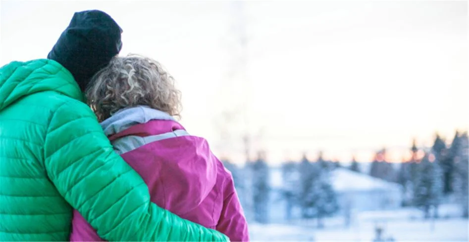 En glad person i vinterkläder med en stickad mössa och tjock jacka står i ett snöigt landskap vid Skellefteå camping. Det ser ut att vara kallt men stämningen är varm och inbjudande.