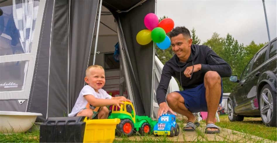 En familj njuter av fritiden på Skellefteå camping. Ett barn sitter i en bilbarnstol bredvid ett uppställt tält, redo för campingäventyr.