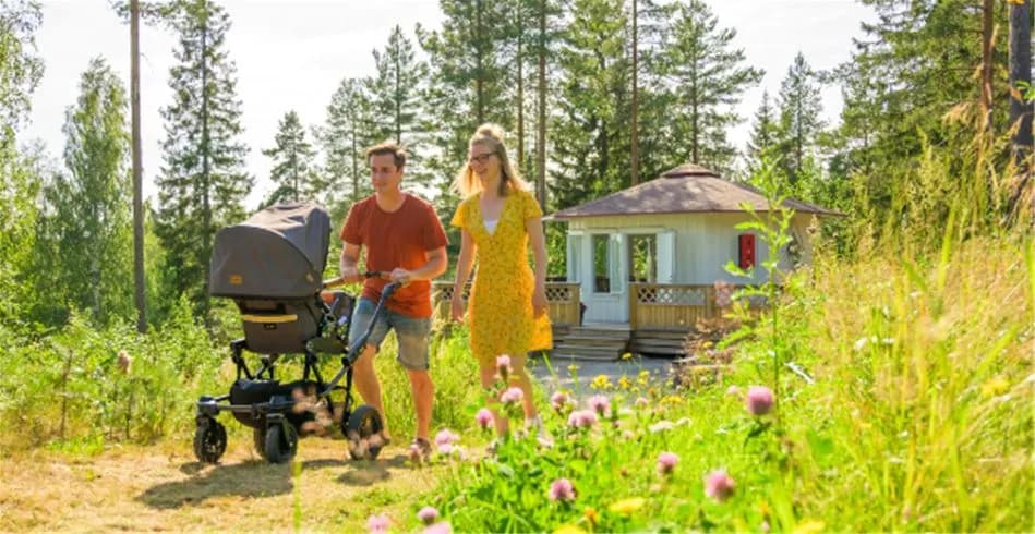 En familj njuter av en solig dag på Skellefteå camping. En vuxen promenerar med ett barn i barnvagn på en grön äng omgiven av tallar, medan ett litet barn leker i närheten, och skapar en avkopplande och familjevänlig atmosfär.