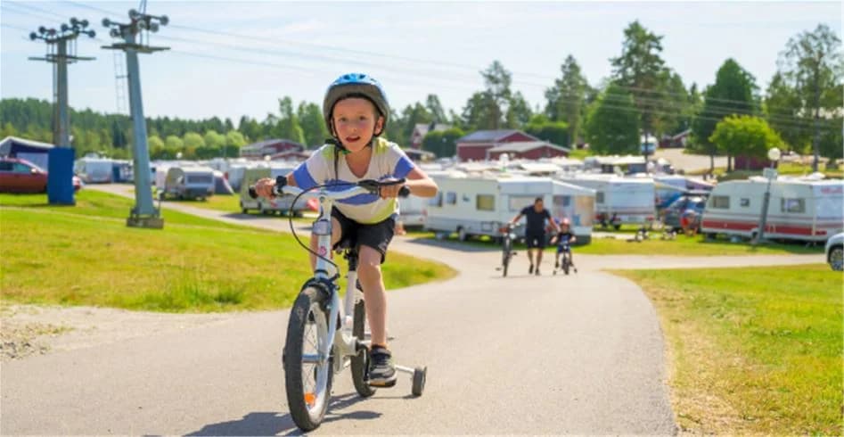 En person bär cykelhjälm och står med en cykel vid Skellefteå Camping på en solig dag, omgiven av grönska.