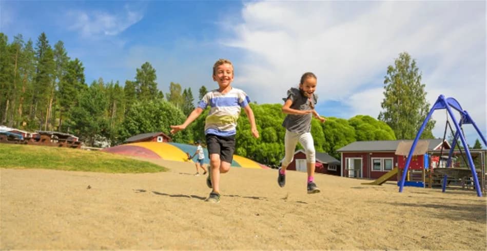 Barn leker på en lekplats utomhus, omgivna av somrig grönska på Skellefteå Camping. Lekutrustningen står i ett öppet och soligt område, vilket lockar till avkopplande och roliga semesterminnen.