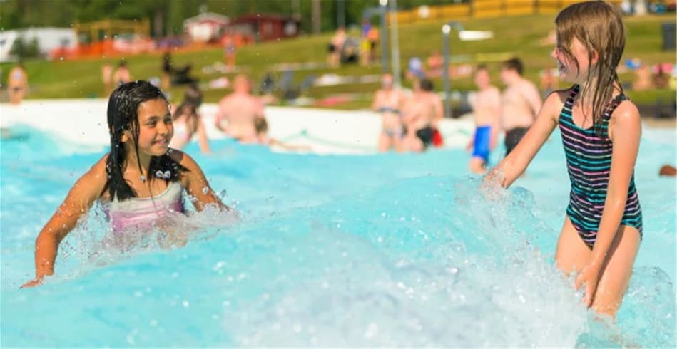 En solig dag på Skellefteå camping med besökare som njuter av sommaraktiviteter vid vattenparken. Barn och vuxna leker, några i baddräkter, och det råder en avslappnad semesterstämning.