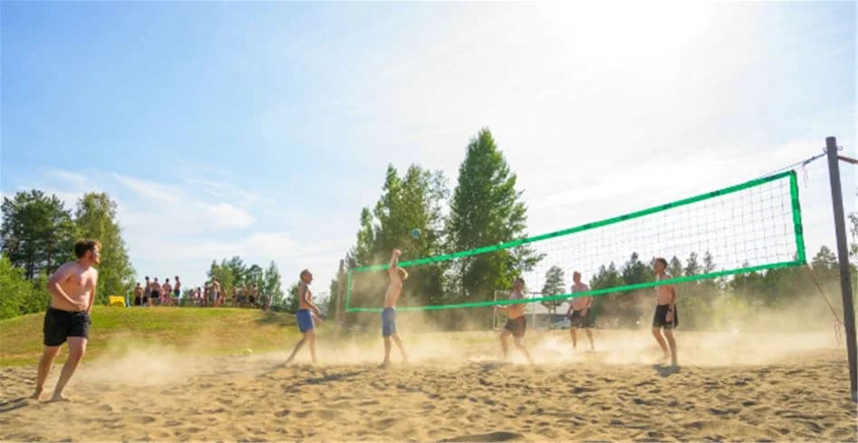Bild av en volleybollplan på Skellefteå camping, där människor spelar beachvolleyboll i solskenet. Bollen är i luften och spelare i shorts syns i bakgrunden.