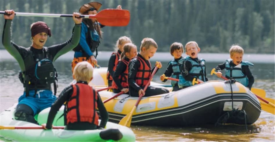 En grupp människor ses i en uppblåsbar båt medan de deltar i vattensporter på en sjö vid Idre Fjäll Camping. Alla bär flytvästar som en säkerhetsåtgärd under deras utomhusäventyr.