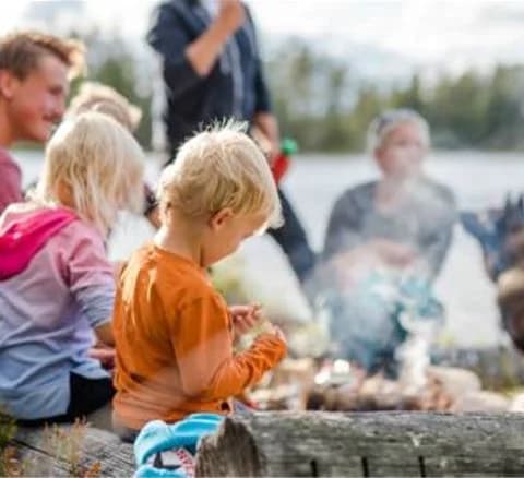 En grupp människor njuter av en lägereld i naturen vid Orsa Grönklitt. Ett barn i förgrunden är fascinerat av lågorna, omgivna av grönska och avkopplande atmosfär.