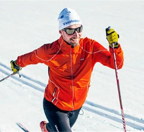 En person klädd i vinterkläder och skidglasögon åker skidor i Orsa Grönklitt, omgiven av snö och vinterlandskap.