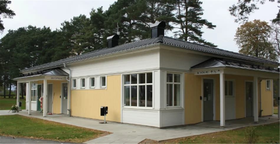 En röd stuga med vita knutar och sadeltak ligger på Tånga Heds camping. Stugan har en välkomnande dörr och är omgiven av grönska, vilket ger en hemtrevlig atmosfär.