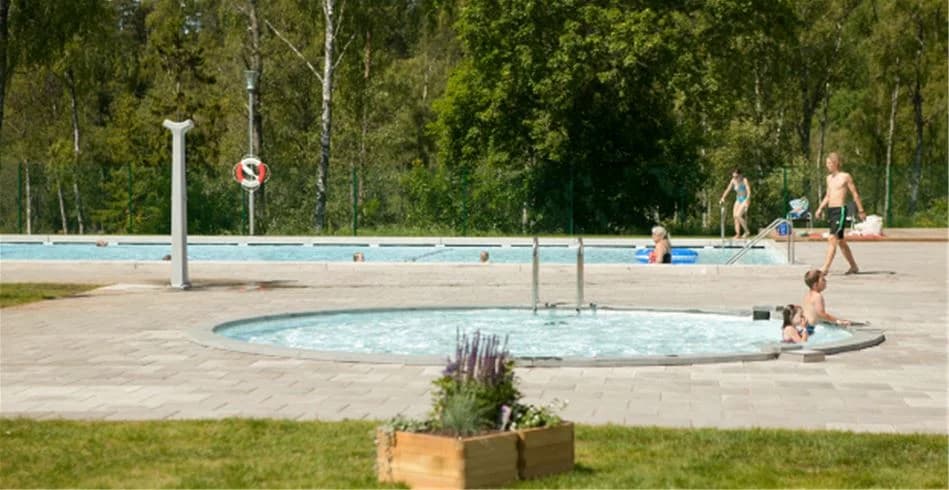 En solig dag vid Tånga Heds Camping med en pool omgiven av utemöbler och glada människor som njuter av ledigheten.