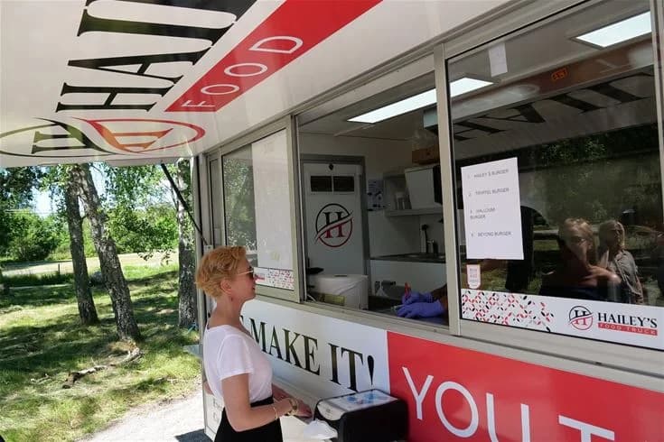 En bild från Björknäs camping visar en matvagn med reklam för snabbmat. Vagnen har en färgglad logotyp och erbjuder mat som hamburgare och pommes frites. I bakgrunden syns naturen.