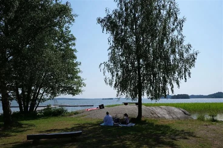 En solig sommardag vid Björknäs camping med gröna gräsmattor, sandstrand och hav i bakgrunden. Människor njuter av fritidsaktiviteter och avkoppling nära kusten.