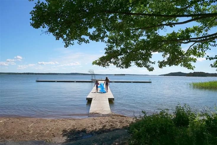 En solig sommardag vid Björknäs Camping med utsikt över ett stilla vatten. Sandstranden leder till en brygga som sträcker sig ut i sjön, omgiven av grönska och kustlinjer.