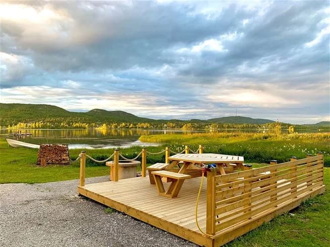 En bild av Överhörnäs camping med en vidsträckt utsikt över ett landskap med kullar och en sjö i bakgrunden. Ett picknickbord står vid stranden under en blå himmel med moln och berg i fjärran.
