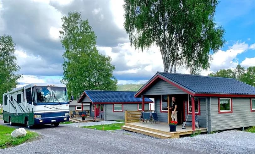 En campingplats med flera stugor och byggnader omgivna av gröna ytor. Parkerade fordon, inklusive husbilar, syns i området.