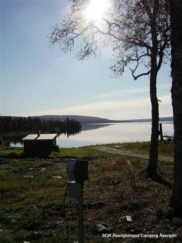 Vy över Åkersjöstrand camping med en glittrande sjö i förgrunden, omgiven av grönskande naturlandskap. Lätt molnig himmel breder ut sig vid horisonten och skapar en lugn och fridfull atmosfär.