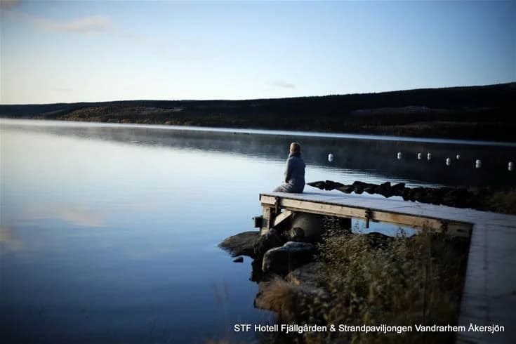 En stilla sjö med en vid horisont, omgivet av kustlandskap och strand. Flera personer vistas vid vattenkanten, njuter av den naturnära miljön på Åkersjöstrand camping.