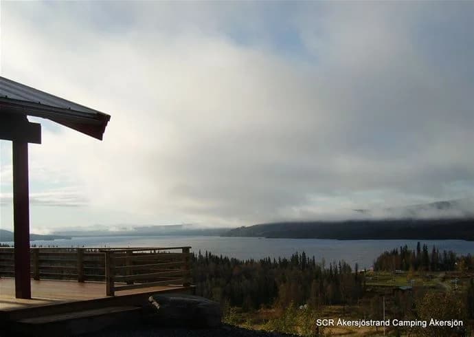 En vy över Åkersjöstrand camping vid sjökanten med moln på himlen, horisontlinje över vattnet och en kustlinje i bakgrunden, fångar känslan av en stilla morgon eller kväll.